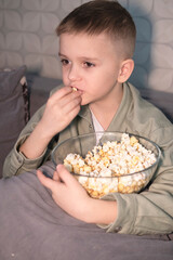 An excited boy is eating popcorn and watching TV with interest. Child is sitting and watching cartoons or movies. Lifestyle. Concept of human emotions. Vertical.