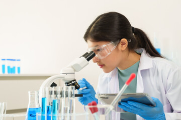 Teenage caucasian girl in lab coat safety goggles and gloves using microscope and holding tablet...