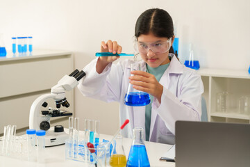 Teenage Caucasian girl in white science coat wearing safety goggles pouring blue liquid from test...
