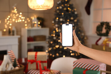 Female hand with mobile phone against glowing Christmas tree and gift boxes at home