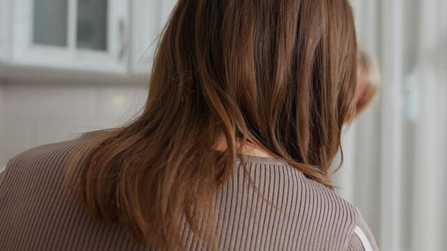 Close-up rear view of young woman with long brown hair holding book while attentively watching her aunt cook in bright kitchen, aunt with short blonde hair is focused on food preparation