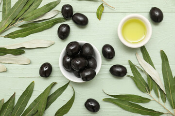 Bowls of oil and tasty black olives with leaves on green wooden background