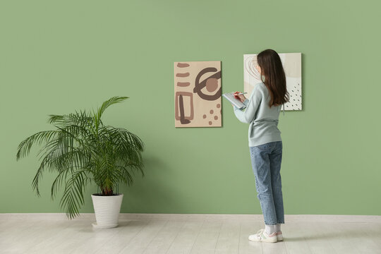 Young woman writing in notebook near paintings on green wall at art gallery exhibition