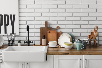 Wooden counter with ceramic sink, clean dishes and kitchen utensils near light brick wall