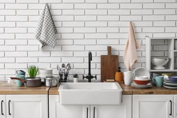 Wooden kitchen counter with sink, clean dishes and towels hanging on light brick wall