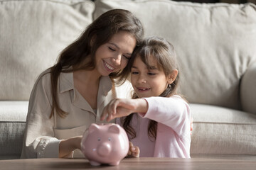 Saving for future. Happy smiling millennial mom little daughter junior schoolgirl hug in living room throwing cash money coins into piggybank. Financial knowledge bank deposit rational economy concept
