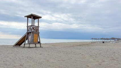 Lifeguard tower on sandy empty beach with overcast cloudy sky and sea view. Wooden lifeguard tower. Serene coastal scene with ocean in the background. Safety concept