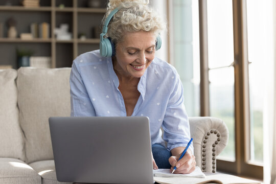 Engaged online learning. Smiling elderly lady in wireless headset remote student at third age university watch virtual lecture webinar on notebook from home office take brief notes in paper copybook