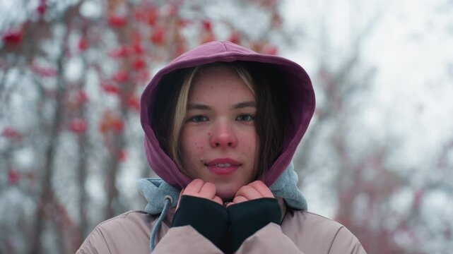 young lady smiling slightly at camera, wearing winter jacket with hood, outdoors, blurred background with trees and snow, casual winter portrait, cold weather, urban park setting