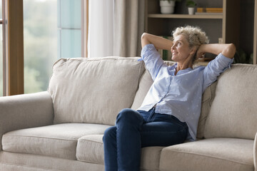 Obraz premium Dreamy and relaxed. Carefree beautiful older age woman in stylish casual wear sit on cozy sofa with dreamy smile looking into distance create plans for future visualize goals relax at home. Copy space