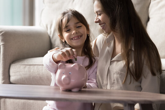 Building financial habits. Cropped close up smiling young mom support happy little kid daughter putting coin to piggy bank. Family finance education domestic economy managing money concept. Copy space