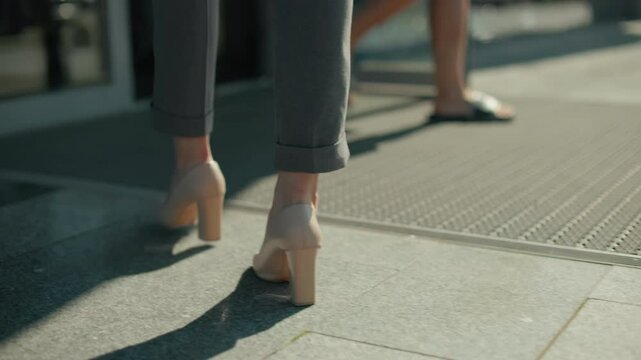Leg view of woman in high heels walking towards open door on sunny day with people exiting building, shadow cast on ground, focus on movement, footwear, and modern urban professional lifestyle