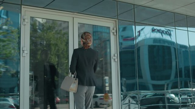 Office worker walks into office building carrying handbag, dressed professionally in black jacket and gray trousers, with glass door reflecting urban street, parked cars, and cityscape on sunny day