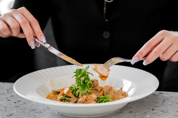 A close-up of a person's hands, cutting into a plate of creamy pasta salad garnished with fresh greens, showcasing the process of enjoying a delicious meal.