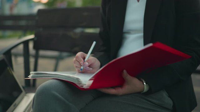 Close up of woman in professional dress reviewing documents in red folder with pen in hand while seated outdoors beside laptop with blurred background featuring benches and greenery