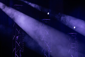 Dramatic purple stage spotlights pierce through misty fog, highlighting metal trusses in a dark...
