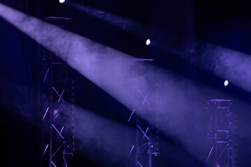 Dramatic purple stage spotlights pierce through misty fog, highlighting metal trusses in a dark theater setting. Strong atmosphere and lighting effects.