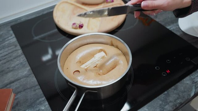 Woman in plaid shirt drops butter into steaming pap as it melts and sinks, electric stove in modern kitchen with wooden tray and dry flowers in background