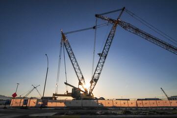 Fototapeta premium Large crawler cranes with extended lattice booms operate at a commercial construction site at sunrise, silhouetted against the sky with safety fencing and signage in the foreground