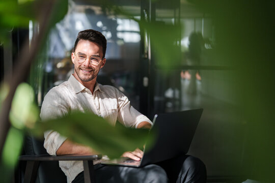 Young Male Professional With Glasses Working On Laptop In Contemporary Office Surrounded By Natural Plants