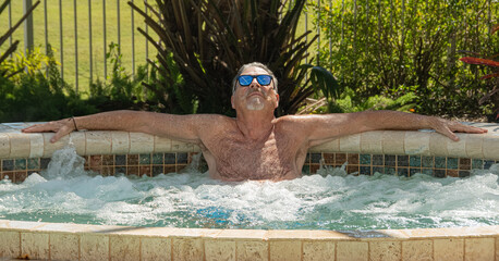 Senior citizen sitting in an outdoor jacuzzi in a tropical setting wearing sunglasses