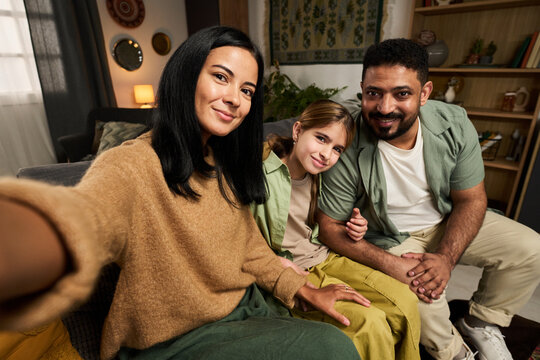 Multiracial family smiling while taking selfie on living room sofa. All members are wearing casual clothing and appear happy and relaxed