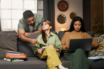 Father discussing math concepts with daughter while another family member focuses on laptop work,...