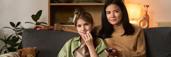 Mother and daughter sitting on sofa, enjoying snacks while watching TV. Surrounding them are cozy home elements, including a lamp and decor behind