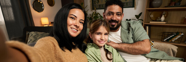 Portrait of smiling family of three sitting on a couch in a cozy living room, capturing a joyful moment together, all looking directly at the camera, creating warm and loving atmosphere