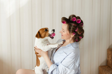 Beautiful young woman wearing pajamas with pink hair curlers holds a cute Jack Russell Terrier dog...