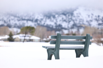 A green bench in the foreground with snow and a blurred house and mountain in the background.  A horizontal landscape presentation of a bleak winter day at the golf course.