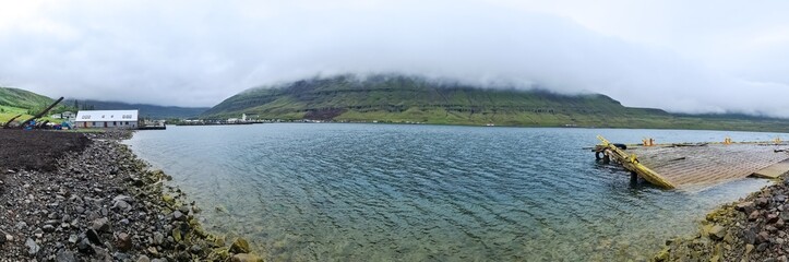 Panoramic view of Seydisfjordur, Iceland, featuring a damaged pier and mountains partially covered by clouds