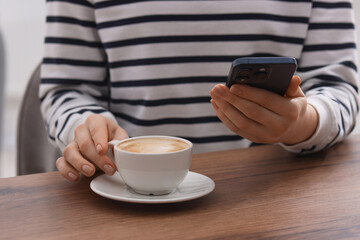 Woman using smartphone during coffee break at wooden table indoors, closeup