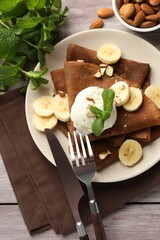 Delicious chocolate crepes with banana, nuts, mint and scoop of ice cream on wooden table, flat lay