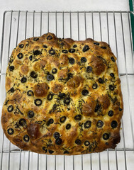 Warm focaccia with black olives rests on a cooling rack, showcasing its golden crust and herbal...