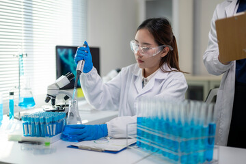 A woman in a lab coat is holding a pipette and looking at a microscope