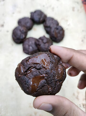 A hand holds a round piece of chocolate cookie dough above a tray lined with multiple cookie mounds, each waiting to be baked. The kitchen exudes a warm, inviting baking atmosphere
