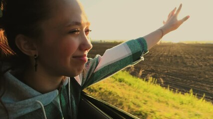 Traveling woman in car stretches hand out of car window, catching fair wind, rays of golden sun. Happy female traveler rides in car, enjoys road trip out of town, watches views of agricultural fields.