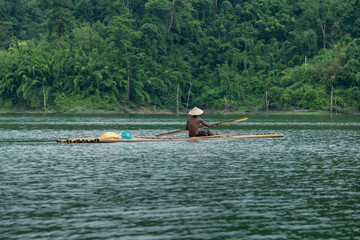 Man paddles a bamboo raft across a serene lake, surrounded by the lush greenery of a tropical rainforest.