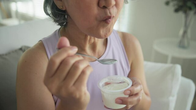 Asia adult people middle age happy old woman eat greek yogurt cup weight loss protein gut health care diet detox sit at home sofa elder aging asian aged female. Fresh medical food relief meal enjoy.