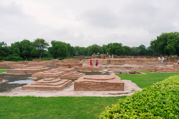 Women walk through ancient brick foundations at Sarnath, Varanasi, surrounded by lush greenery and remnants of Buddhist monasteries.