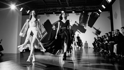 Black and white video of a fashion runway show, captured from a low angle, highlighting models in dramatic lighting and bold abstract backdrop. - Powered by Adobe