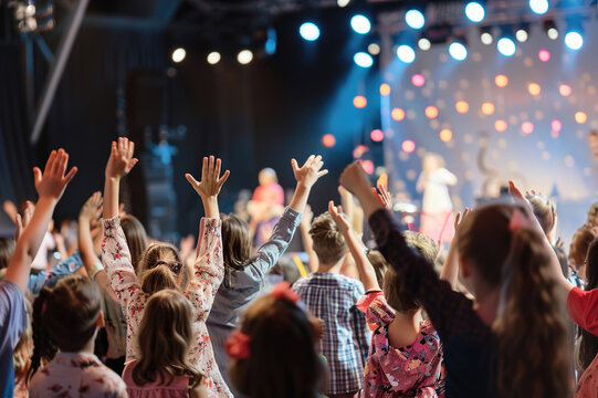 Kids on stage performing, vibrant costumes, colorful backdrop, audience clapping, and bright lights