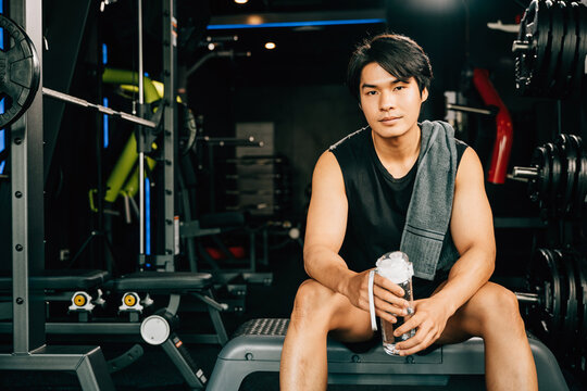 Portrait of Asian young man trained body in black sportswear sitting for rest holding water or protine in fitness gym, Weight training, healthy lifestyle concept