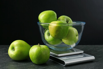 Electronic kitchen scale with bowl of green apples on dark textured table against black background, closeup
