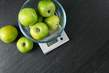 Electronic kitchen scale with bowl of green apples on dark textured table, flat lay. Space for text