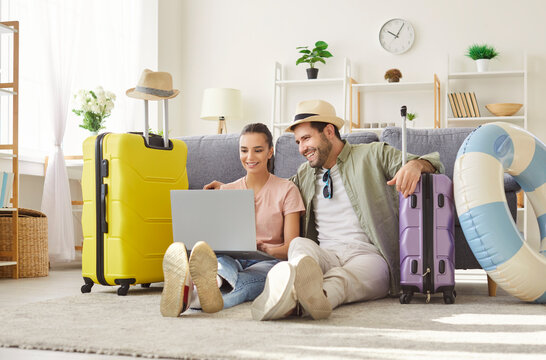 Happy smiling couple travelers sitting on floor with suitcases at home booking tickets online via laptop. Young man and woman wearing beach hat choosing tour for holiday trip. Vacation concept.