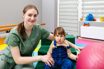 Happy child sitting with his physiotherapist during session of physical rehabilitation and exercise. Physical rehabilitation of children and early intervention