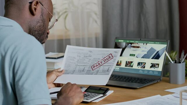Medium rear shot of African American male homeowner sitting at desk, holding overdue utilities invoice, calculating living costs and writing down numbers in journal, while managing household budget