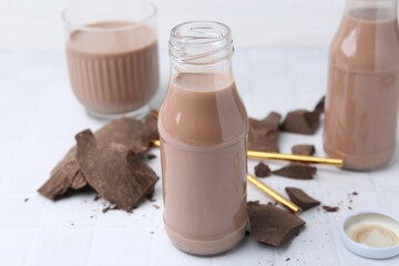 Tasty chocolate milk in glass bottles and pieces of chocolate on white tiled table, closeup